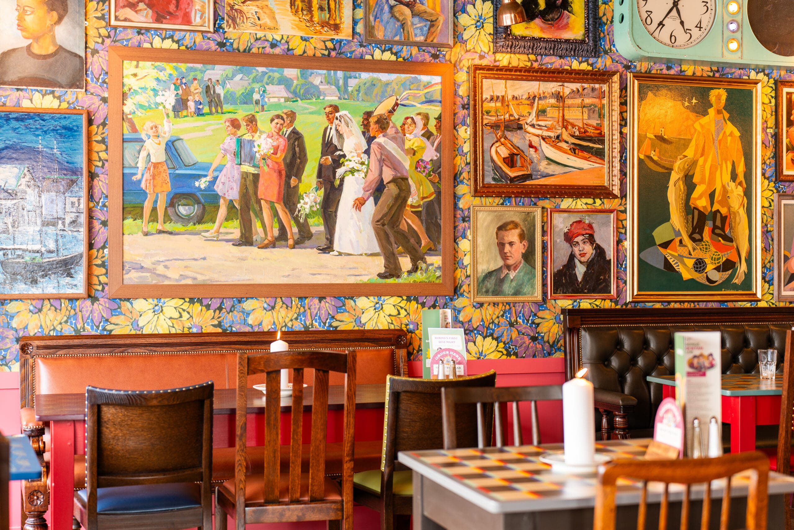 A vibrant café interior with mismatched chairs and tables, bright floral wallpaper, and walls covered in colorful framed paintings depicting people, boats, marinero scenes, and portraits. A menu and candle sit on the front table.