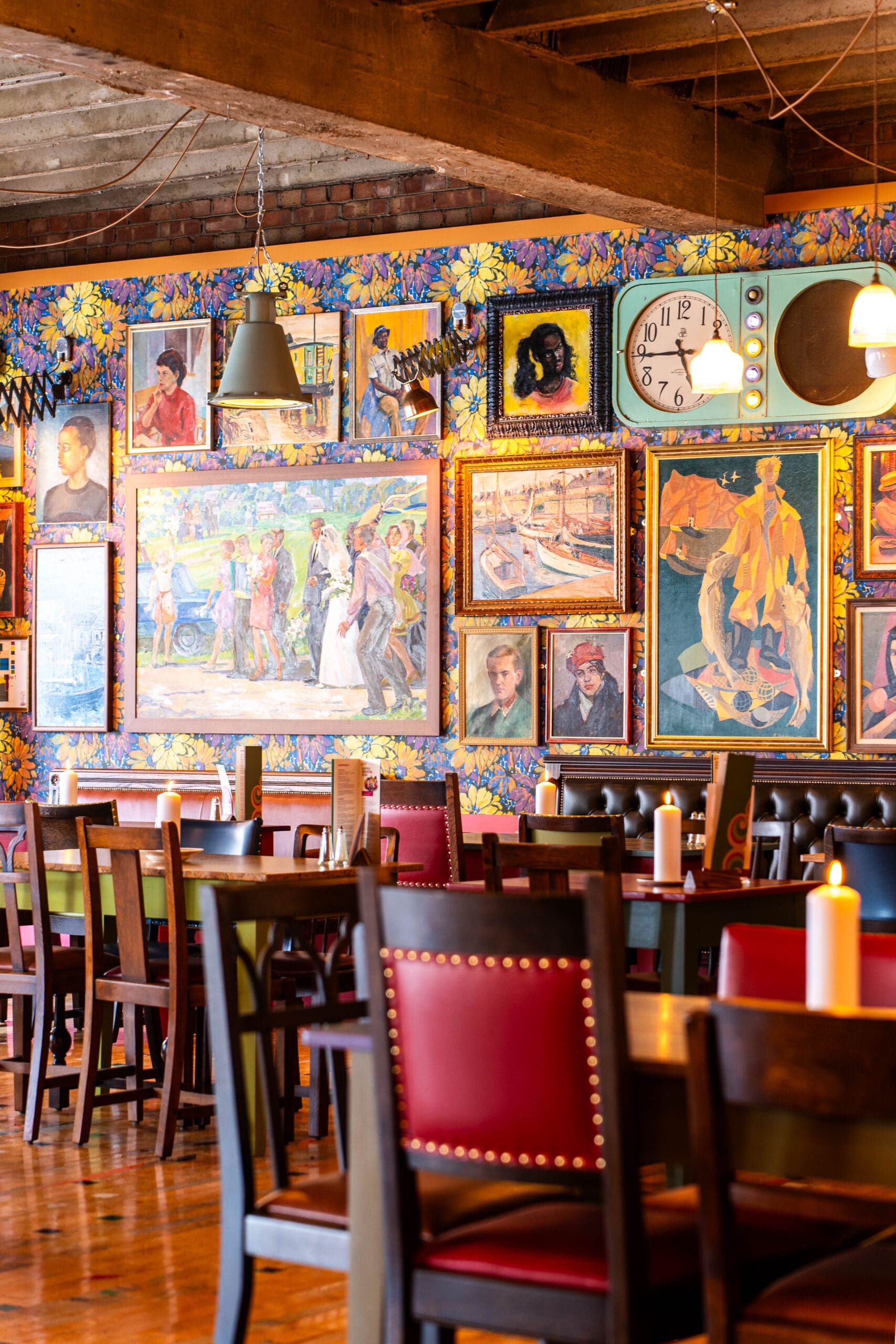 A colorful restaurant interior with wooden tables and chairs, red and green seat cushions, and walls decorated with vibrant paintings, portraits, a retro clock, a vintage TV, and subtle marinero touches against a floral wallpaper background.