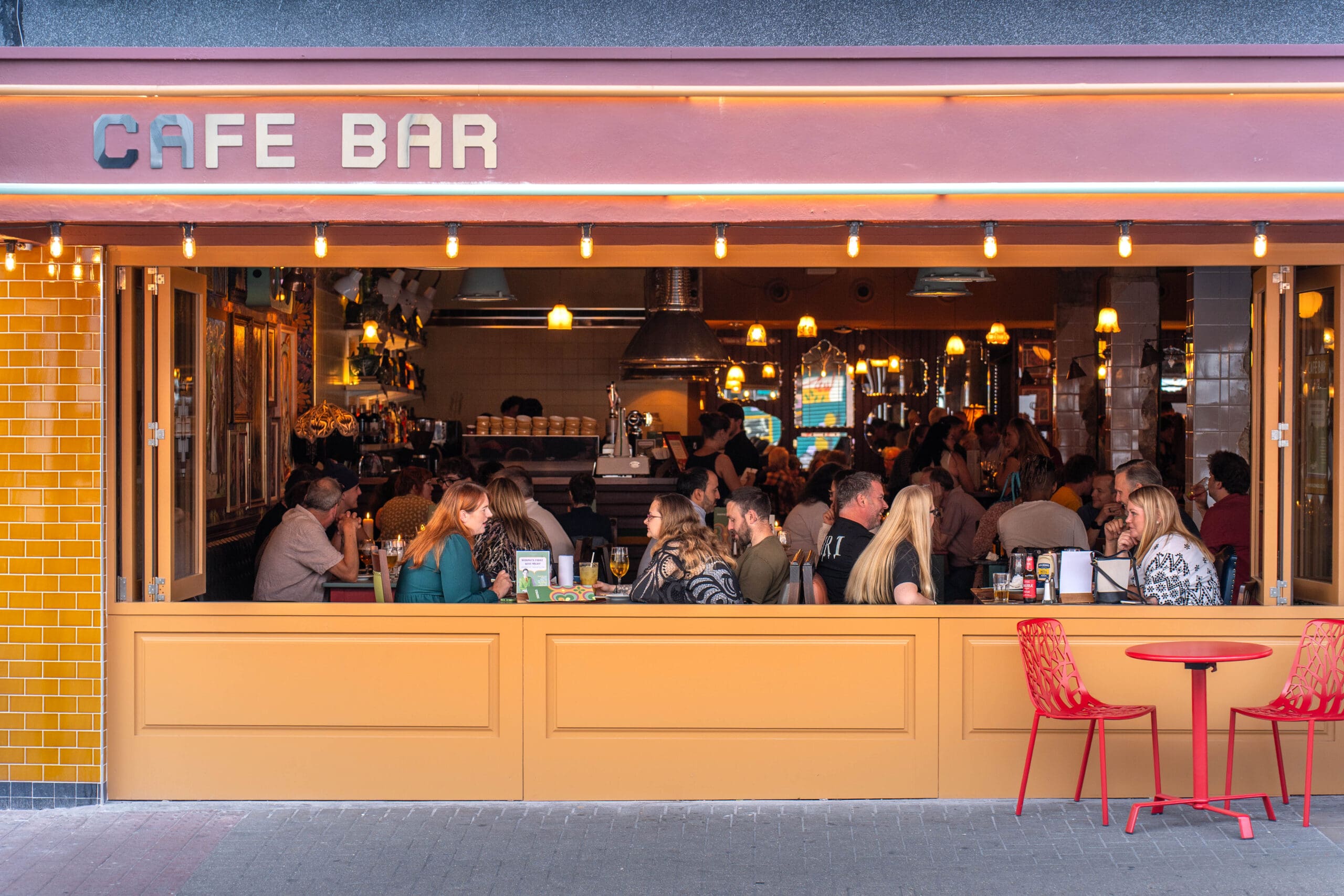 People sit at tables inside a lively, warmly lit café bar with a bright yellow and orange exterior. The open front reveals customers chatting and dining while two empty red chairs await outside, evoking a relaxed marinero ambiance.