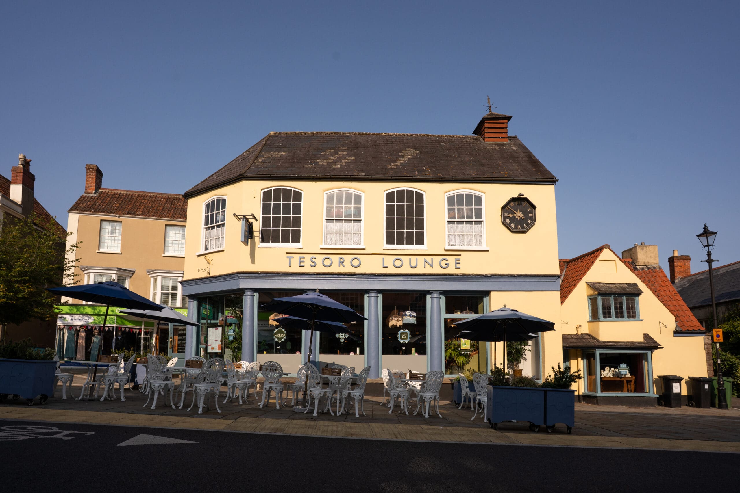 A two-story building with "Tesoro Lounge" signage features large windows and outdoor seating with Bianco tables and chairs under blue umbrellas on a sunny day. A clock adorns the building, with neighboring houses nearby.