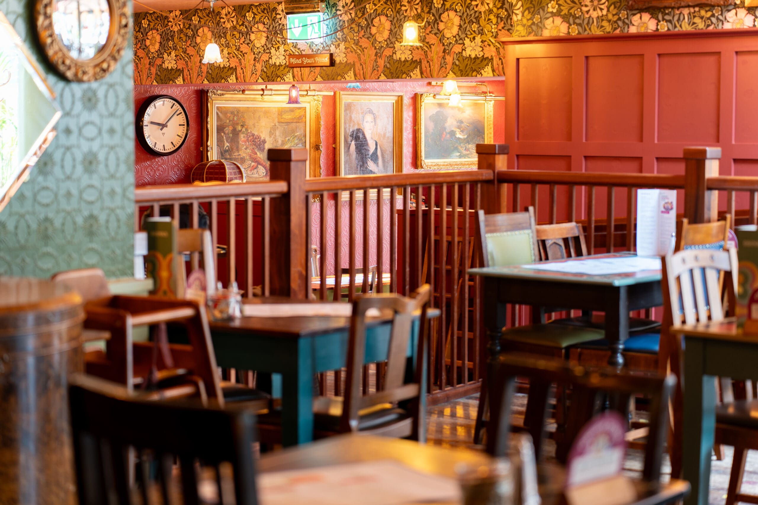 Cozy vintage-style café interior with Bianco wooden tables and chairs, patterned wallpaper, framed paintings, a clock on the wall, and warm, ambient lighting.