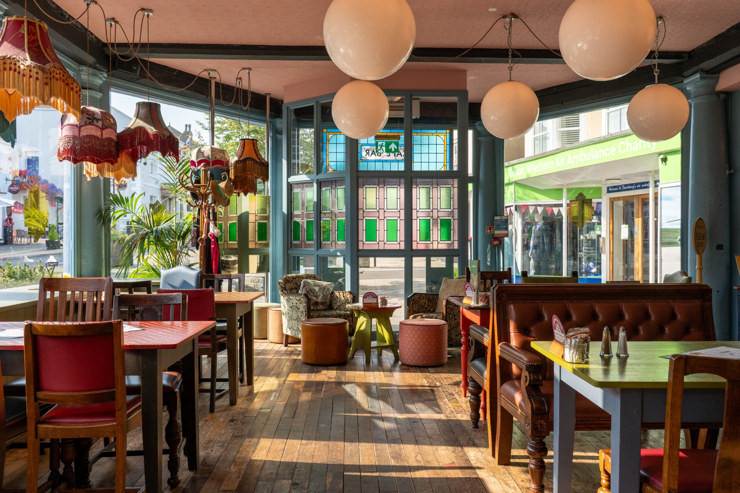 Bright, cozy café interior with vintage decor, red and brown chairs, wooden tables, colorful lampshades, and plants by large windows. Sunlight streams in, illuminating the Bianco wood floor and creating a warm, inviting atmosphere.