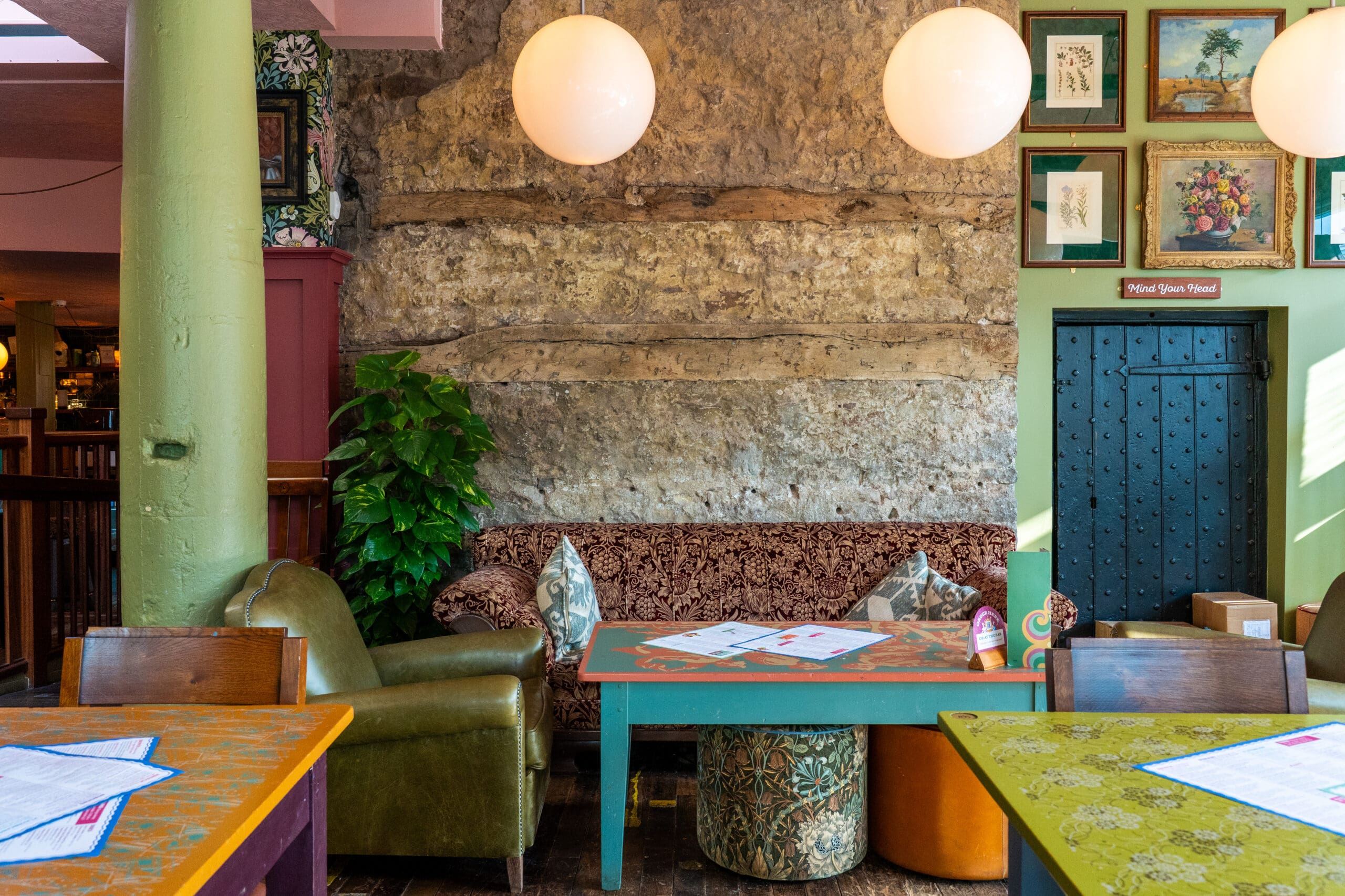A cozy café interior with vintage furniture, Bianco-patterned tables, a green armchair, a floral sofa, round hanging lights, a textured stone wall, framed art, and a green door. Menus are placed on the tables.