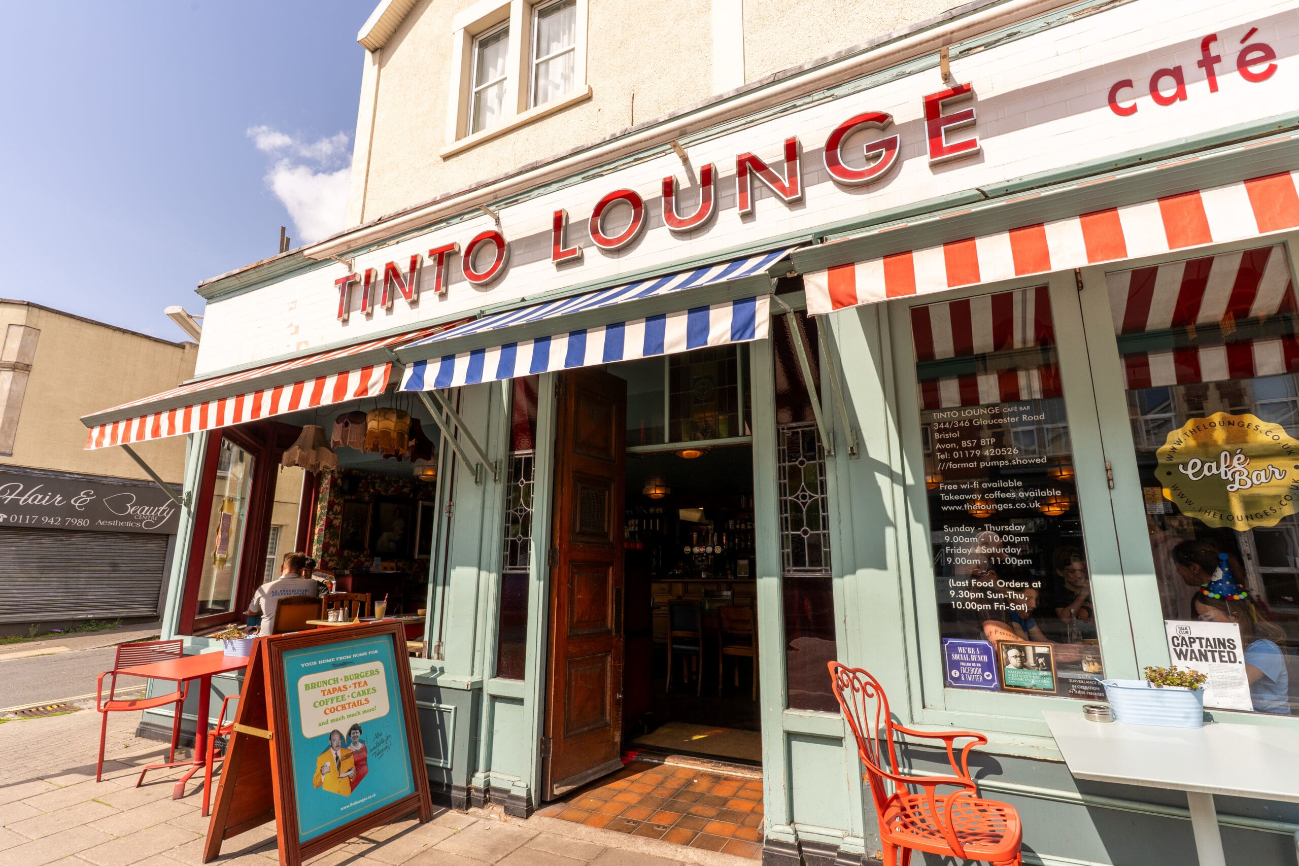 The exterior of Tinto Lounge café, with its signature red and blue striped awning and Tinto signboard, features inviting outdoor seating where a person enjoys a sunny day at a table near the open door.