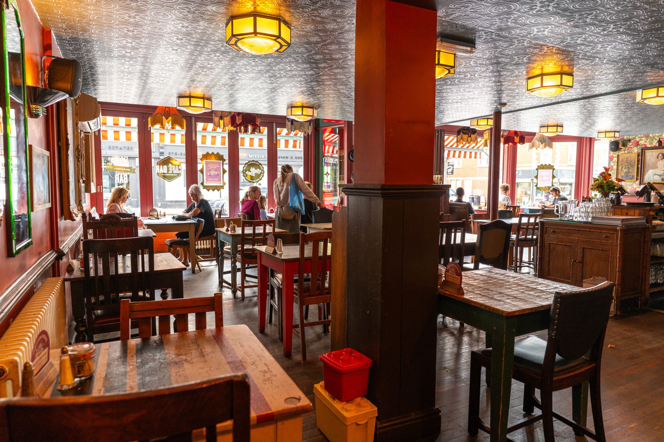 A cozy café interior with wooden tables and chairs, warm lighting, and several people enjoying Tinto near windows decorated with red-and-white striped awnings. The atmosphere appears relaxed and inviting.