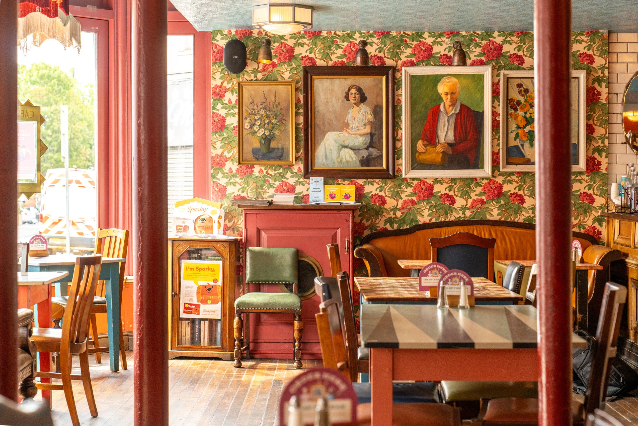 A cozy café interior with floral wallpaper, vintage furniture, and framed paintings—including portraits and flowers—adorns the walls. Wooden tables and chairs fill the warmly lit space, creating a welcoming Tinto atmosphere.
