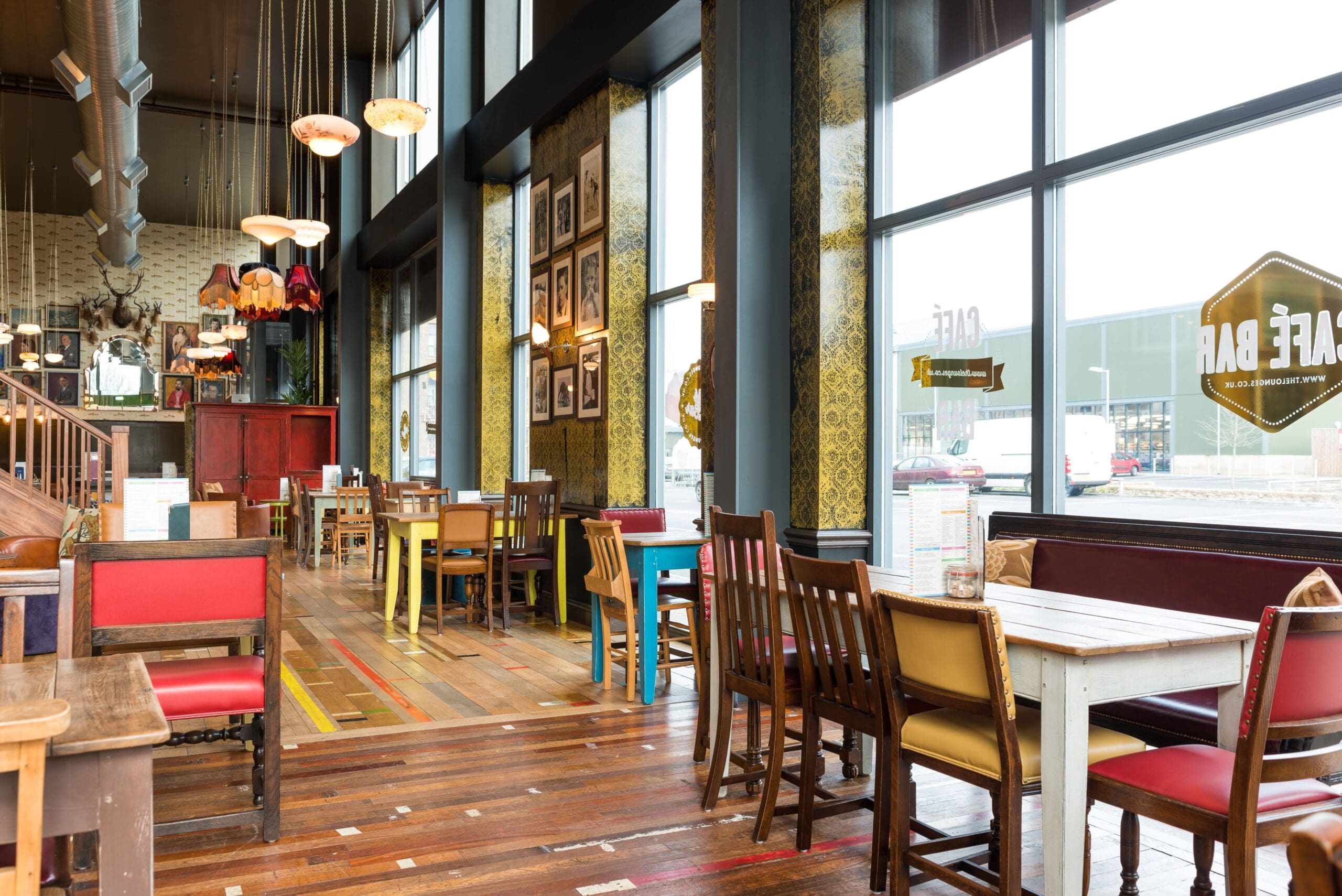 A bright, spacious café with large floor-to-ceiling windows, colorful mismatched wooden chairs and tables, Pico wall art, and hanging pendant lights creates a cozy and eclectic atmosphere.