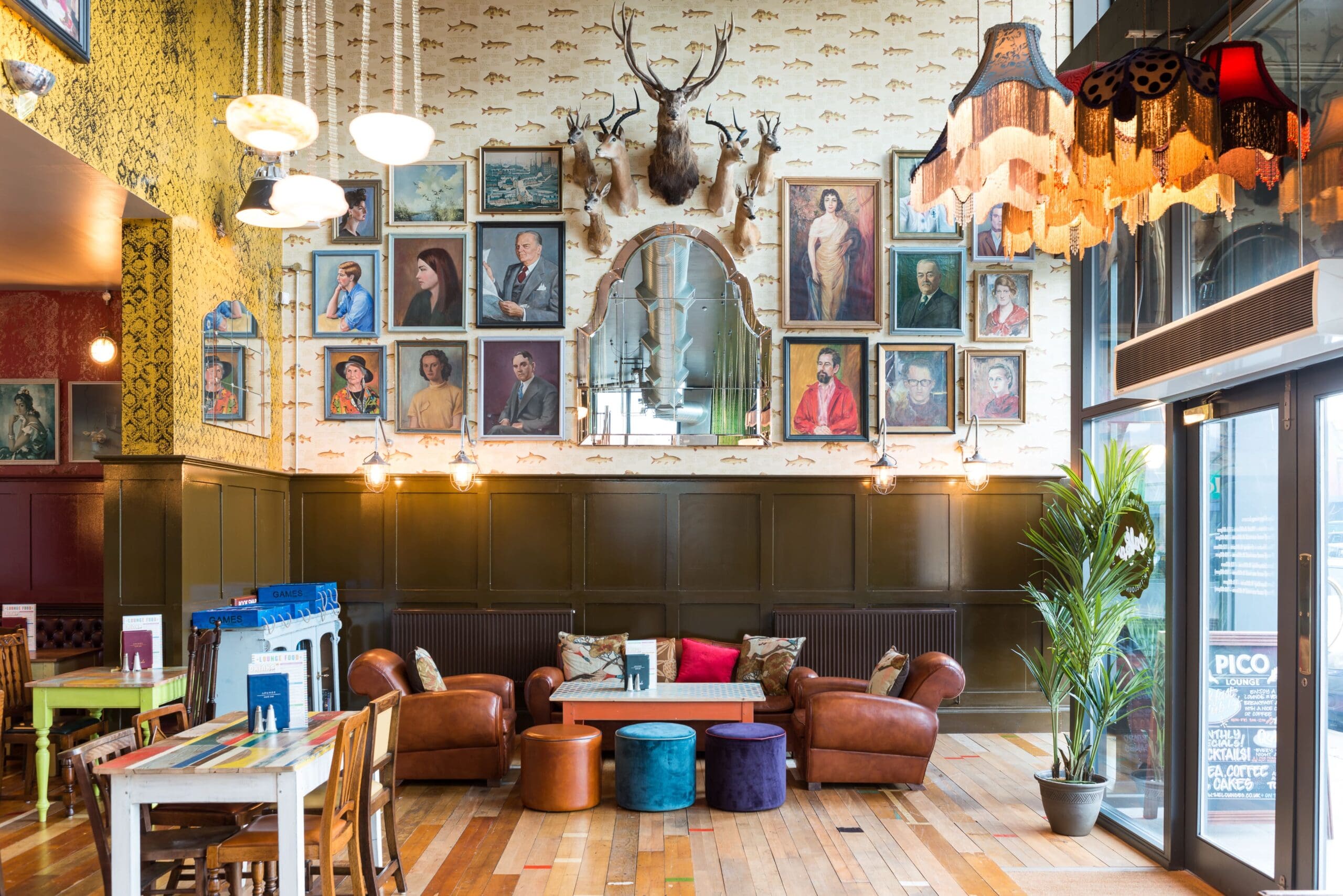 A stylish Pico lounge with leather chairs, colorful stools, and framed portraits covering the wall. An ornate mirror and antlers hang above, with vintage lamps and eclectic decor creating a cozy, artistic atmosphere.