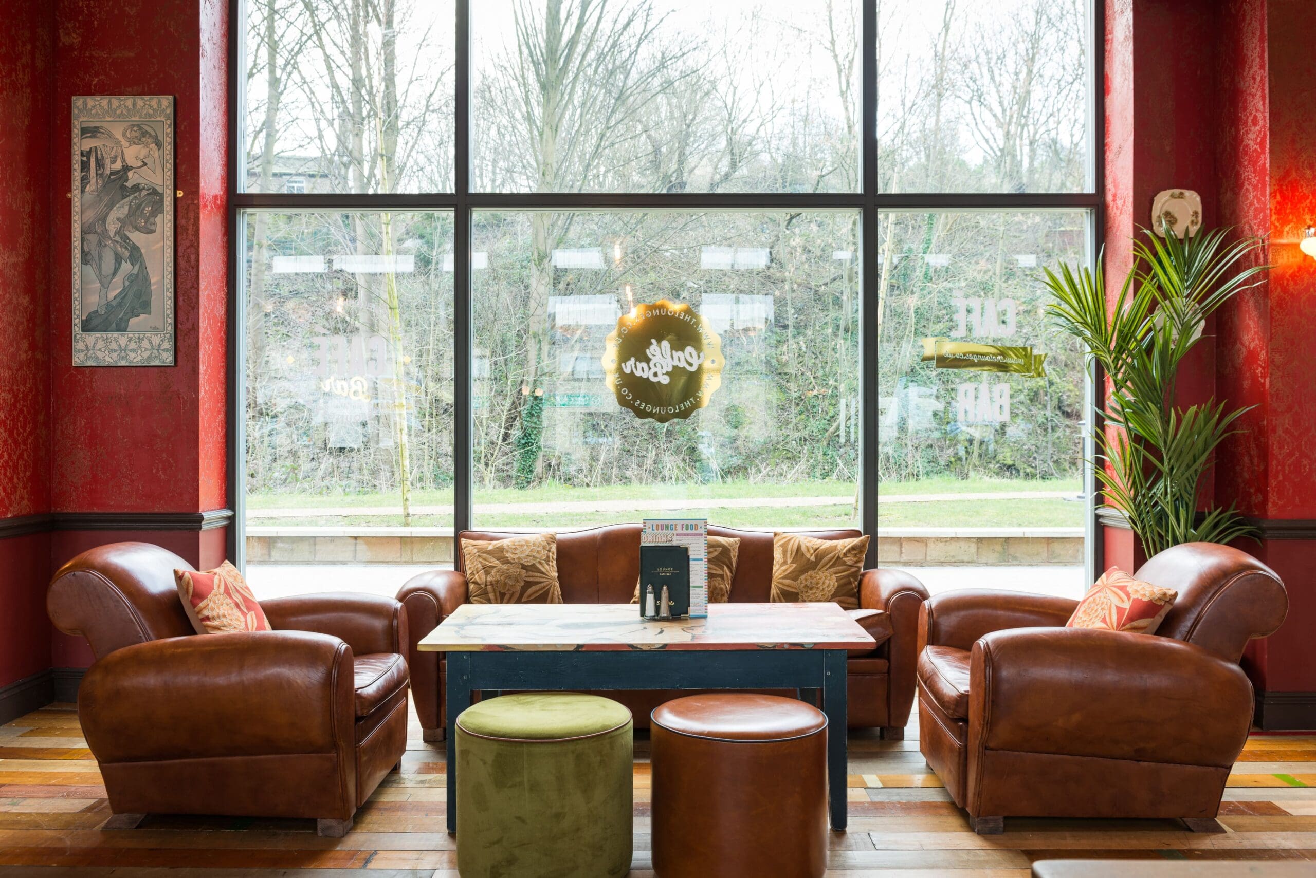Cozy seating area with brown leather armchairs and a sofa around a wooden coffee table, two green ottomans, houseplants, and a large window showing Pico’s cityscape. Warm red walls and a wooden floor add comfort.