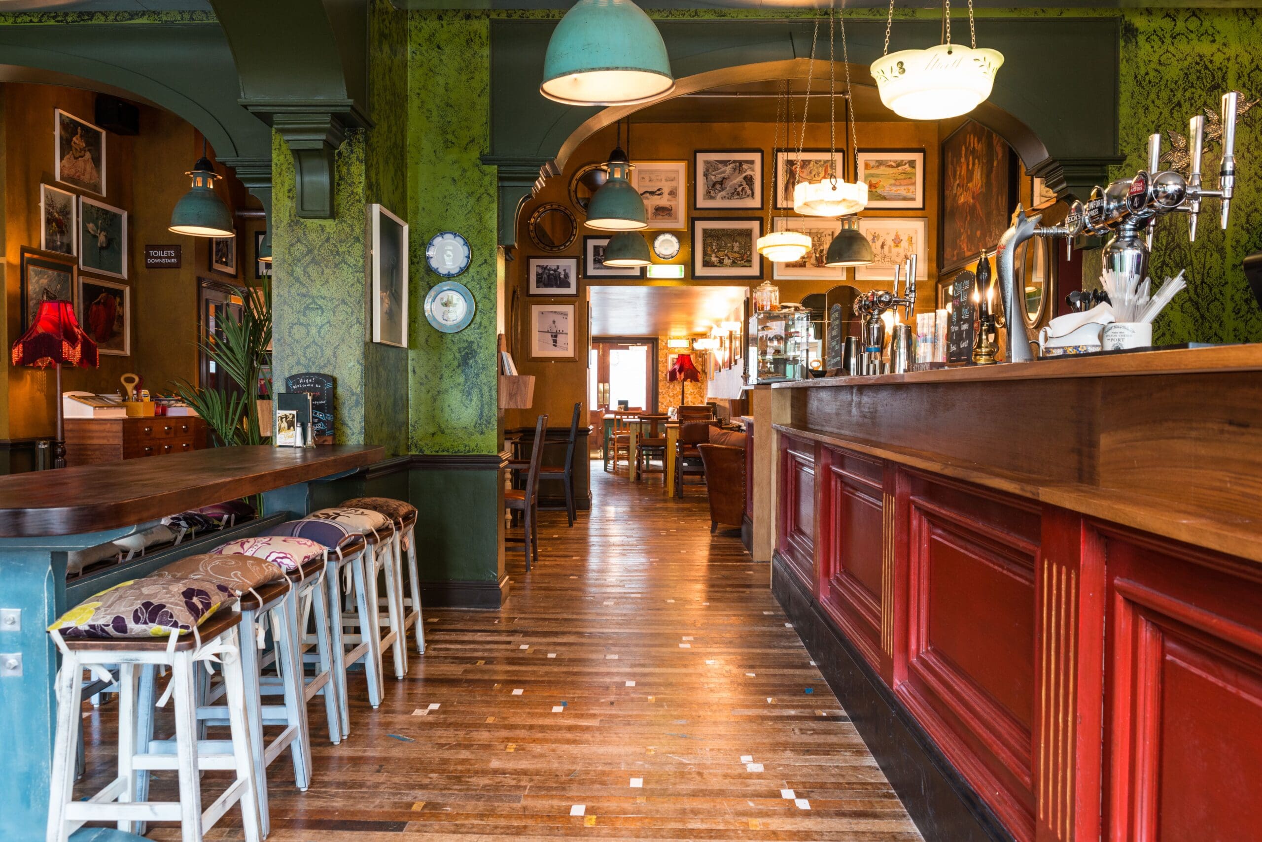 Cozy pub interior with wooden floors, a long red bar, patterned stools, green walls, and vintage light fixtures. Framed pictures and eclectic details create a welcoming Milo vibe in this inviting space.