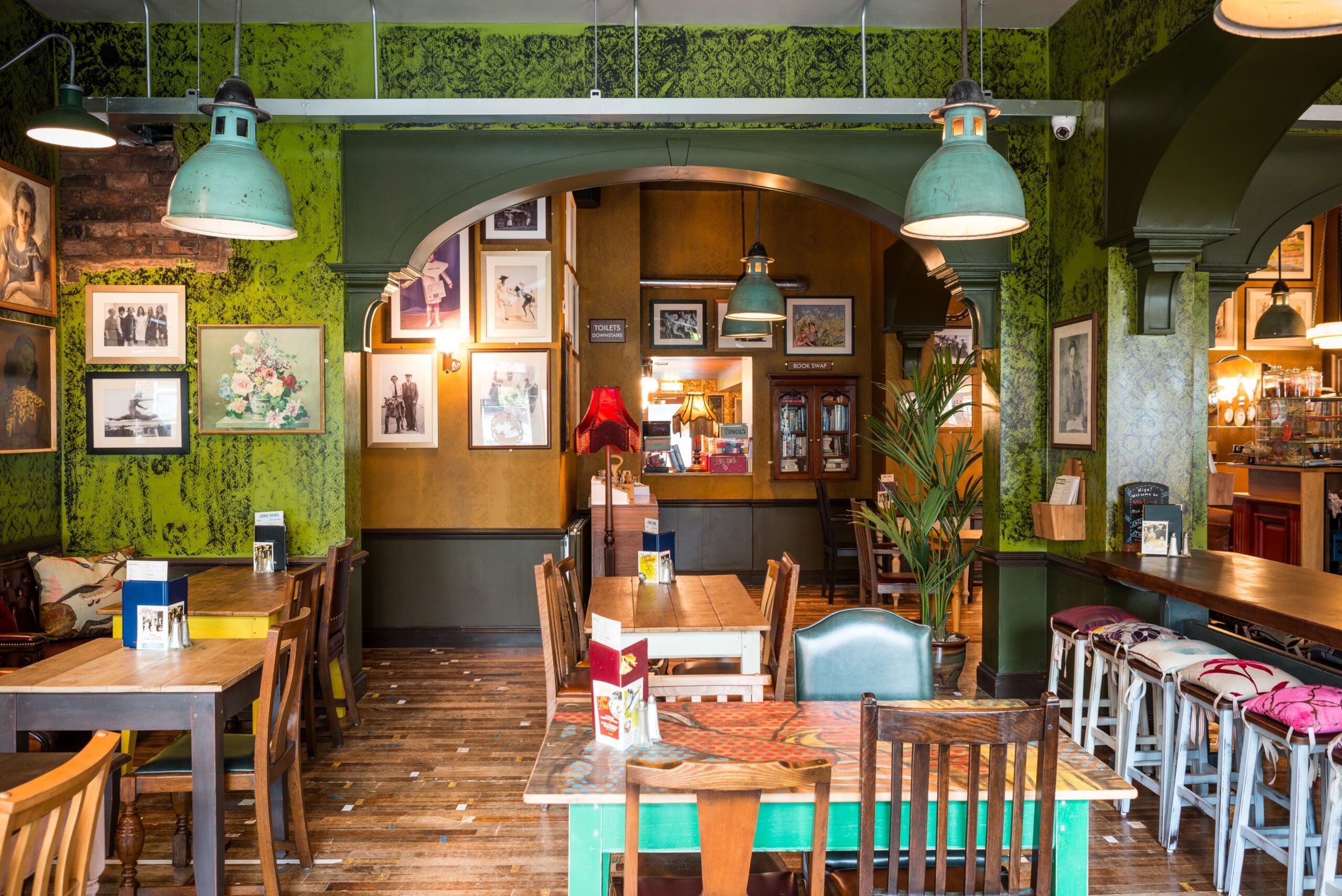 A cozy café interior with mismatched wooden tables and chairs, green patterned wallpaper, framed photos and art on the walls, pendant lights, and a relaxed, eclectic atmosphere—perfect for sipping Milo and unwinding.