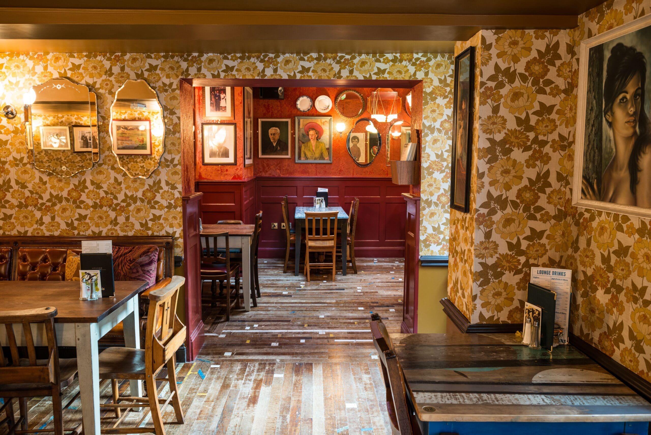 A cozy vintage-style pub with floral wallpaper, mismatched wooden chairs and tables, framed artwork on the walls, and warm ambient lighting. The wooden floor adds to Milo’s inviting and eclectic atmosphere.