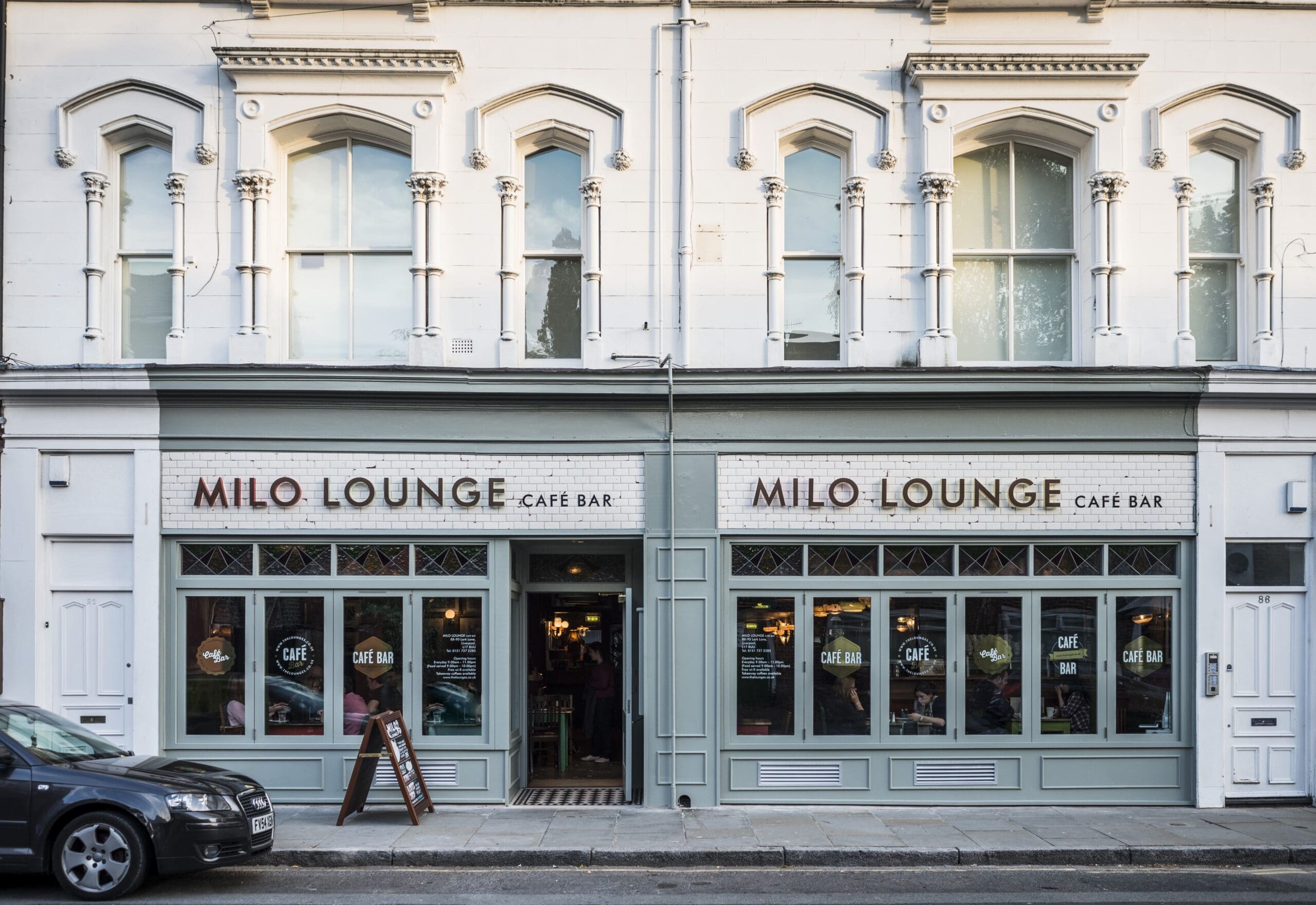 A street view of Milo Lounge Café Bar, with large windows, a light green and white Milo-themed façade, and two visible signs. A car is parked in front, and the welcoming café interior is visible through the entrance.