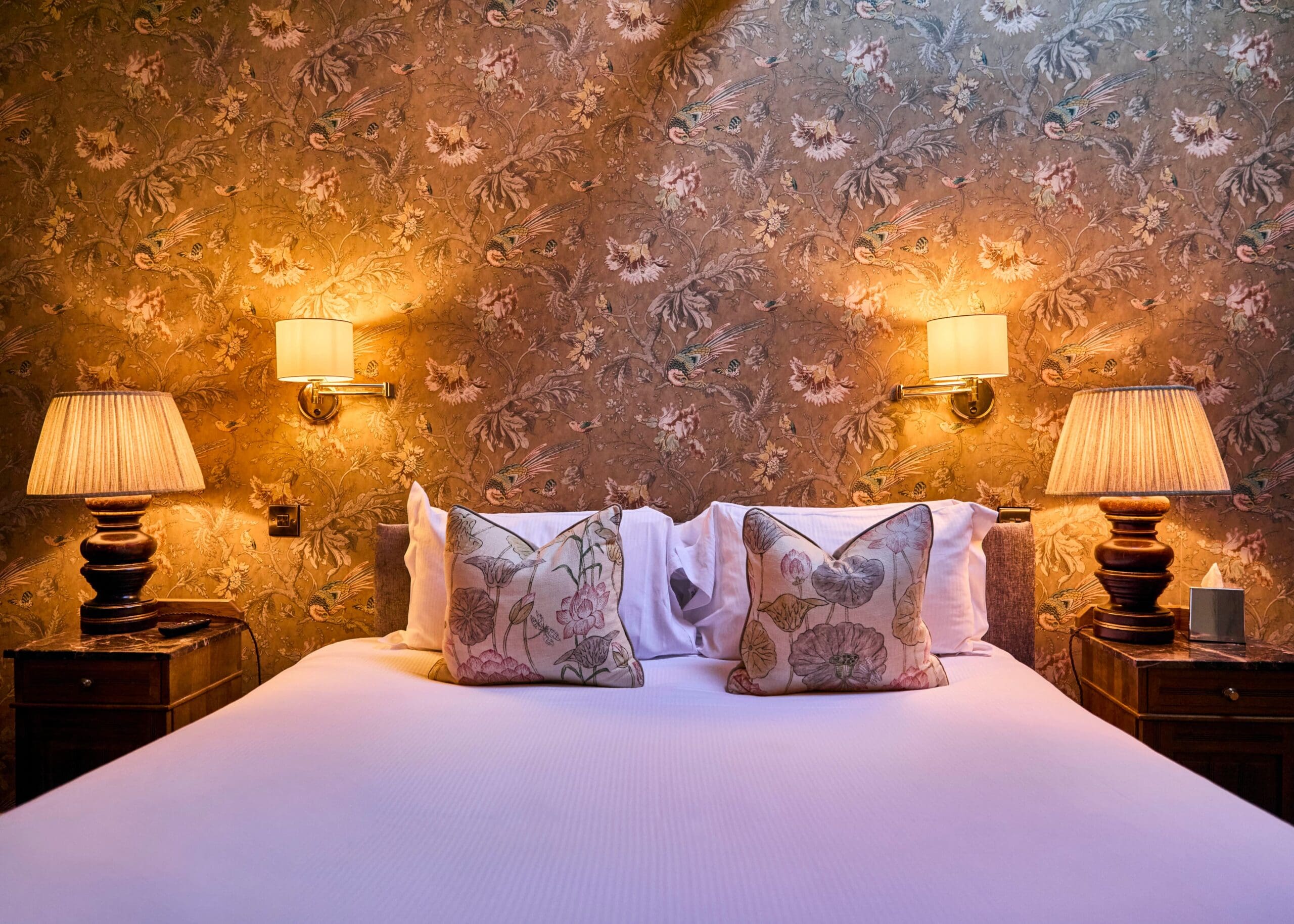 A cozy bedroom with a large bed, two floral pillows, and white bedding. The bed is flanked by wooden nightstands, matching table lamps, and wall sconces. The background features vintage-style floral wallpaper.