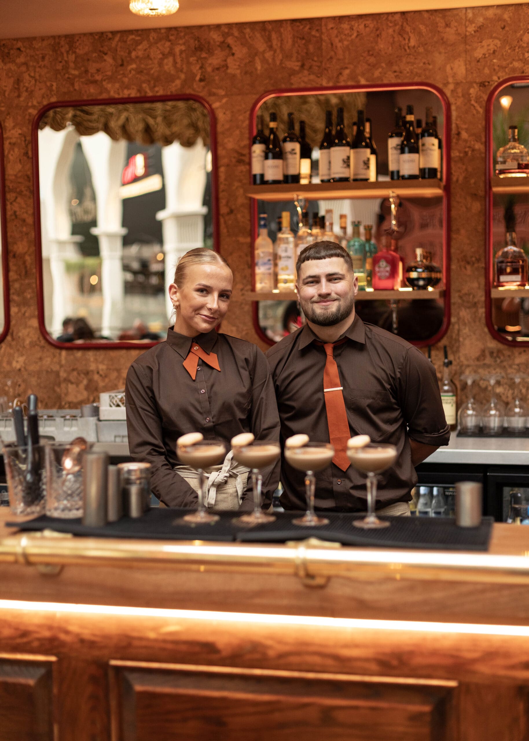 Two bartenders in matching brown shirts and orange ties stand behind a bar lined with four espresso martinis. Shelves with bottles and mirrors are behind them, creating a warm, inviting atmosphere.