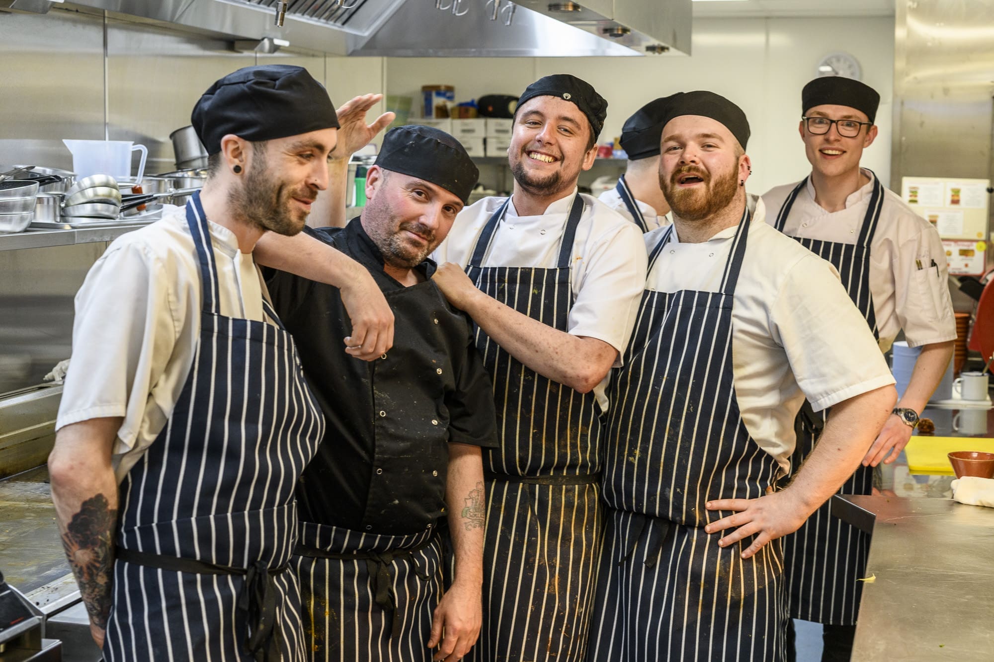Five chefs in striped aprons and black hats smile and pose together in a commercial kitchen, showcasing their rewarding culinary careers amid stainless steel appliances and utensils.