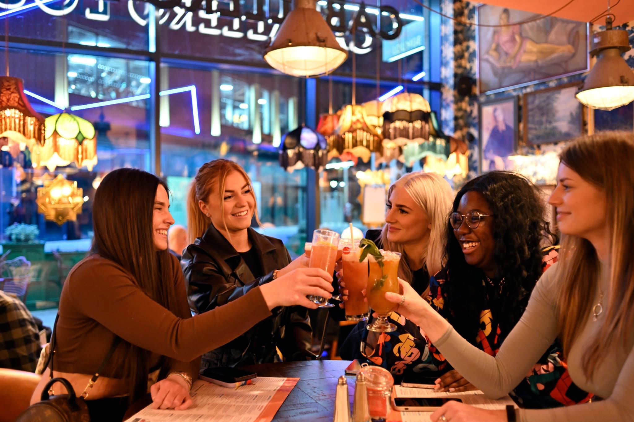 Five women sit around a table at a colorful, modern restaurant, smiling and clinking their vibrant cocktails together in a celebratory toast. Menus and drinks are on the table, and warm lighting fills the space.