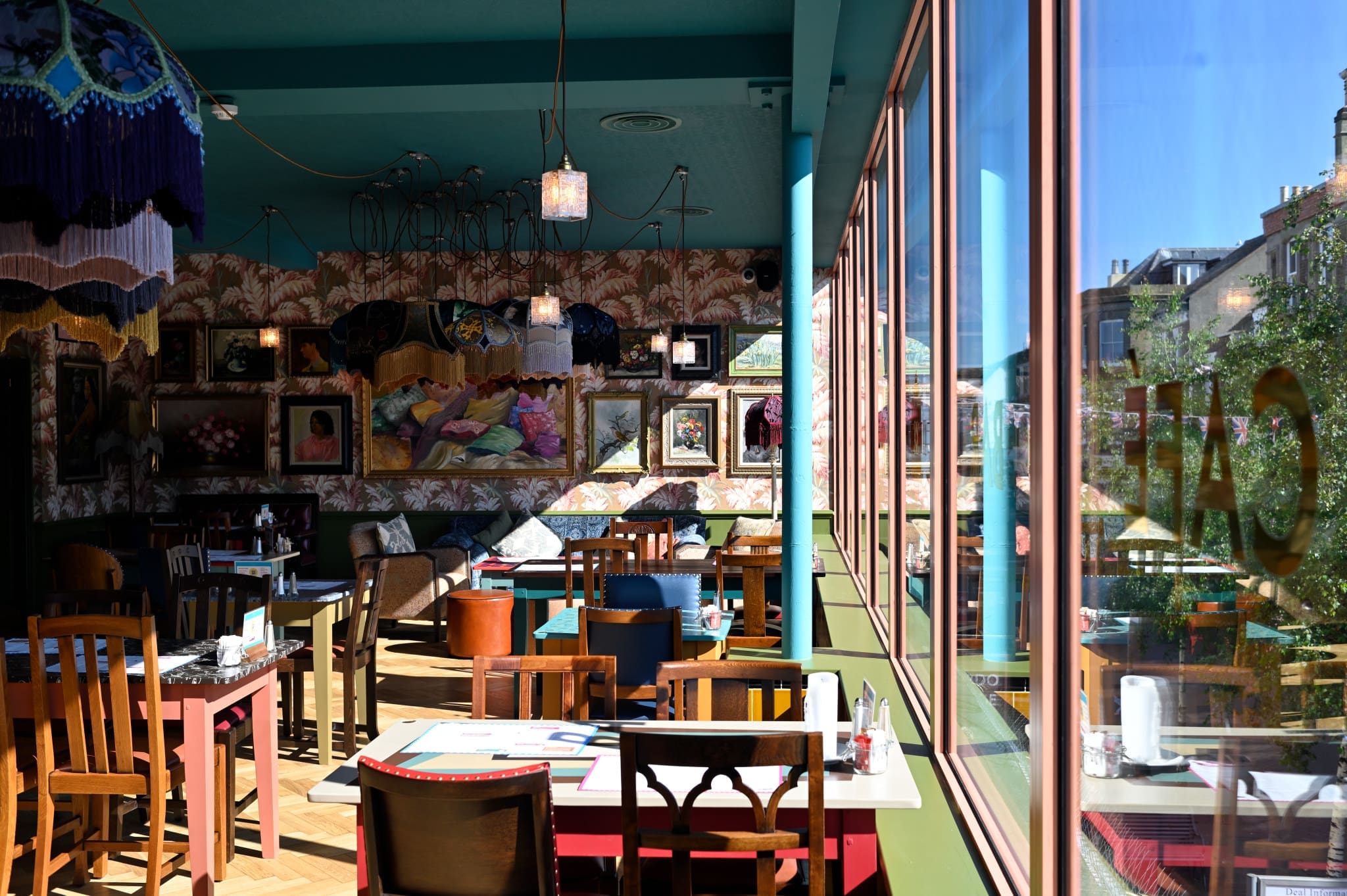 Sunlit café interior featuring eclectic decor, colorful wall art, and mismatched wooden chairs and tables. Large windows on the right let in bright daylight and offer a view of buildings outside—an inviting spot for an Acordo coffee break.