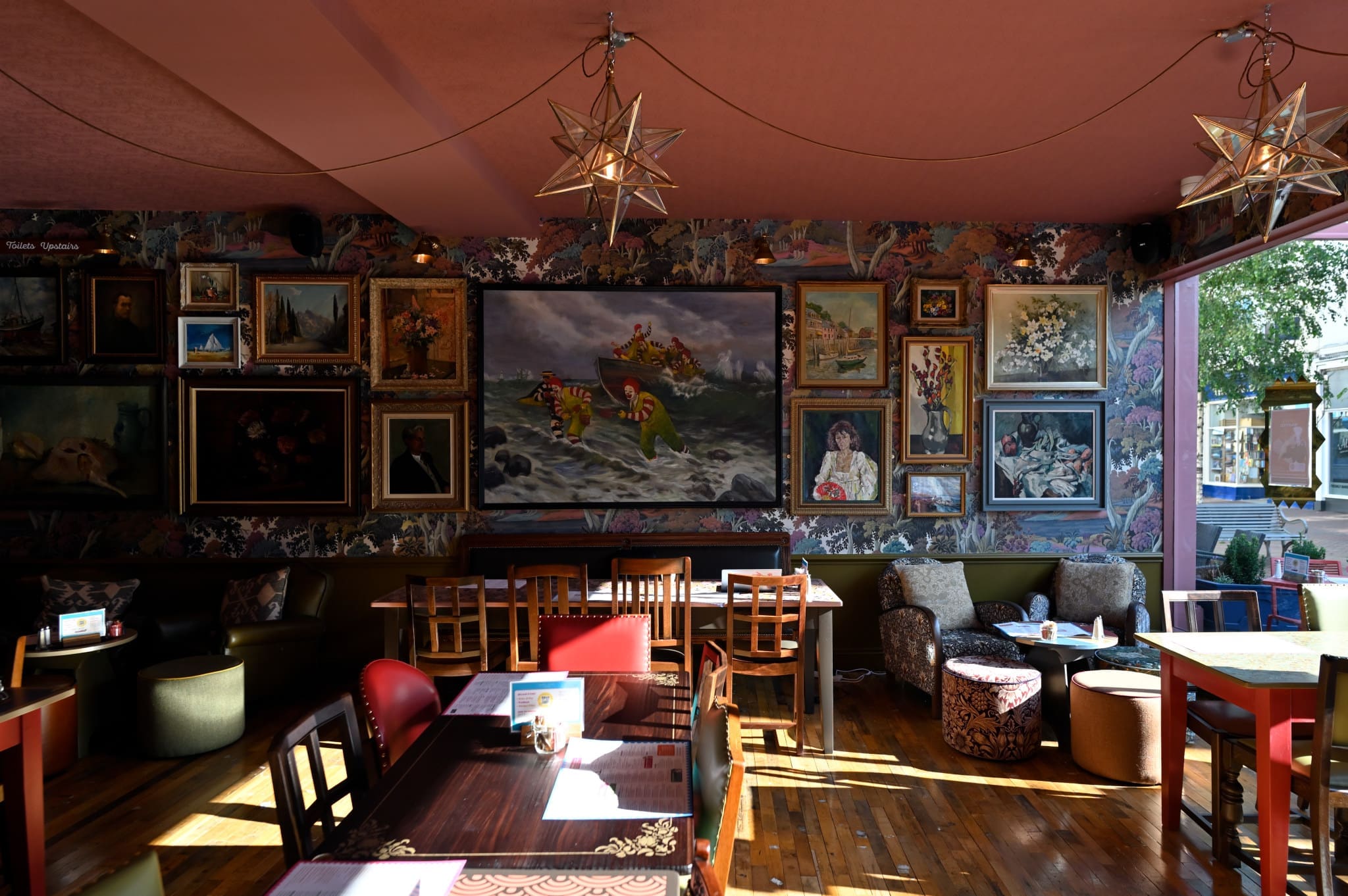 Acordo is a cozy cafe with wooden tables and chairs, floral wallpaper, eclectic framed artwork on the walls, star-shaped hanging lights, and large windows letting in sunlight.