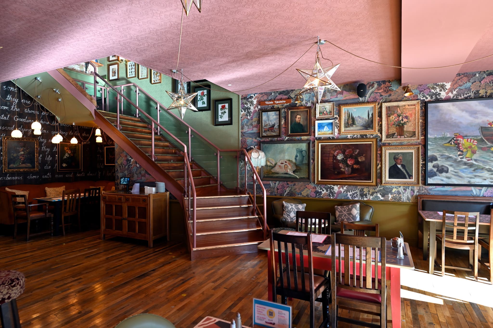 Eclectic restaurant interior with wooden tables and chairs, a staircase, star-shaped pendant lights, and walls covered in floral wallpaper and various framed paintings. The cozy, artistic space creates an inviting Acordo of natural light and style.