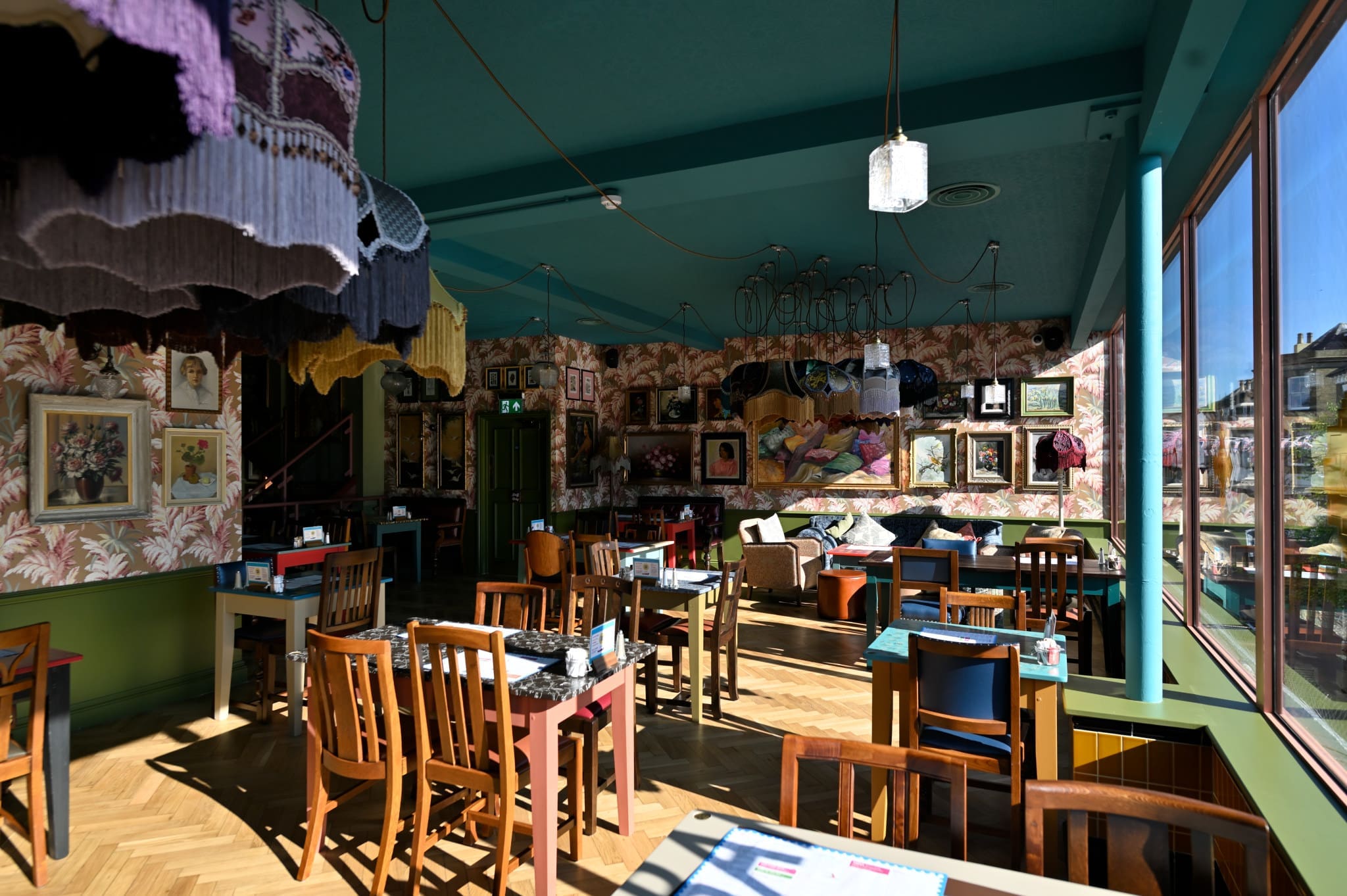 A sunlit, eclectic café interior with mismatched wooden chairs, colorful tables, and patterned wallpaper sets a cozy Acordo vibe. Framed art adorns the walls while large windows and fringed lamps hang from the teal ceiling.