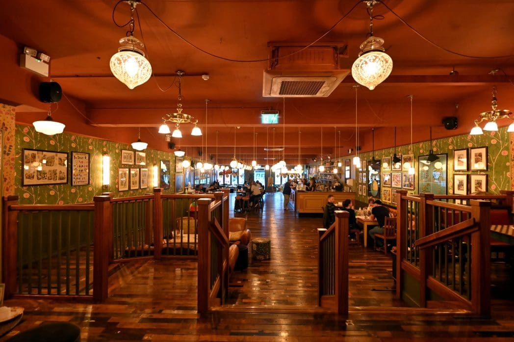 Warmly lit pub interior with wooden floors, vintage-style light fixtures, and patterned wallpaper. Framed pictures and patrons create an inviting Alento vibe, with a central aisle leading to more cozy seating.
