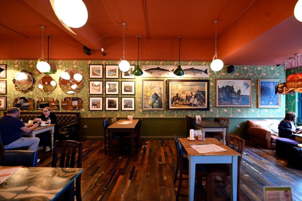 A cozy, eclectic cafe, Alento, features patterned green wallpaper, a colorful ceiling, framed art and mirrors on the walls, wooden floors, and people dining at tables or a couch near the window. Warm hanging lights create an inviting atmosphere.
