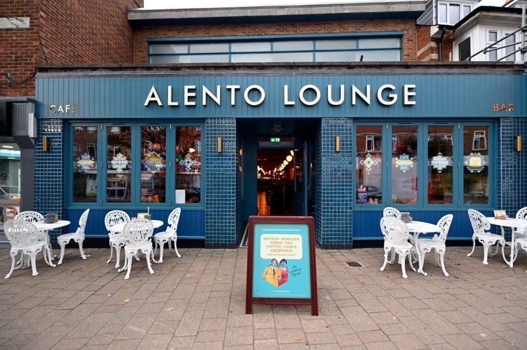 The Alento Lounge café exterior features signature blue tiles, large windows, white outdoor tables and chairs, and a sandwich board sign welcoming guests at the entrance on a paved sidewalk.