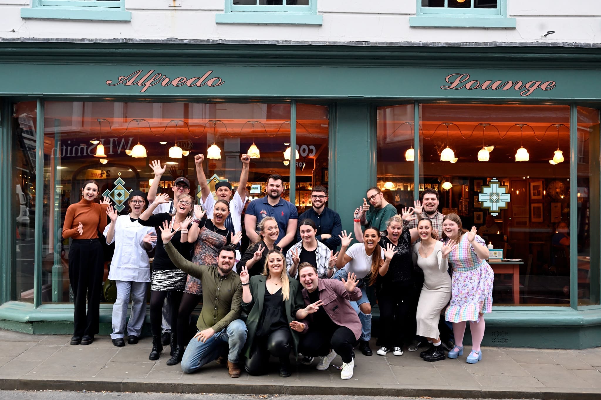 A group of people pose and smile enthusiastically outside Alfredo Lounge, a cafe or restaurant with large windows, Bordo accents, and teal signage. Some have their arms raised, and the group appears joyful and celebratory.