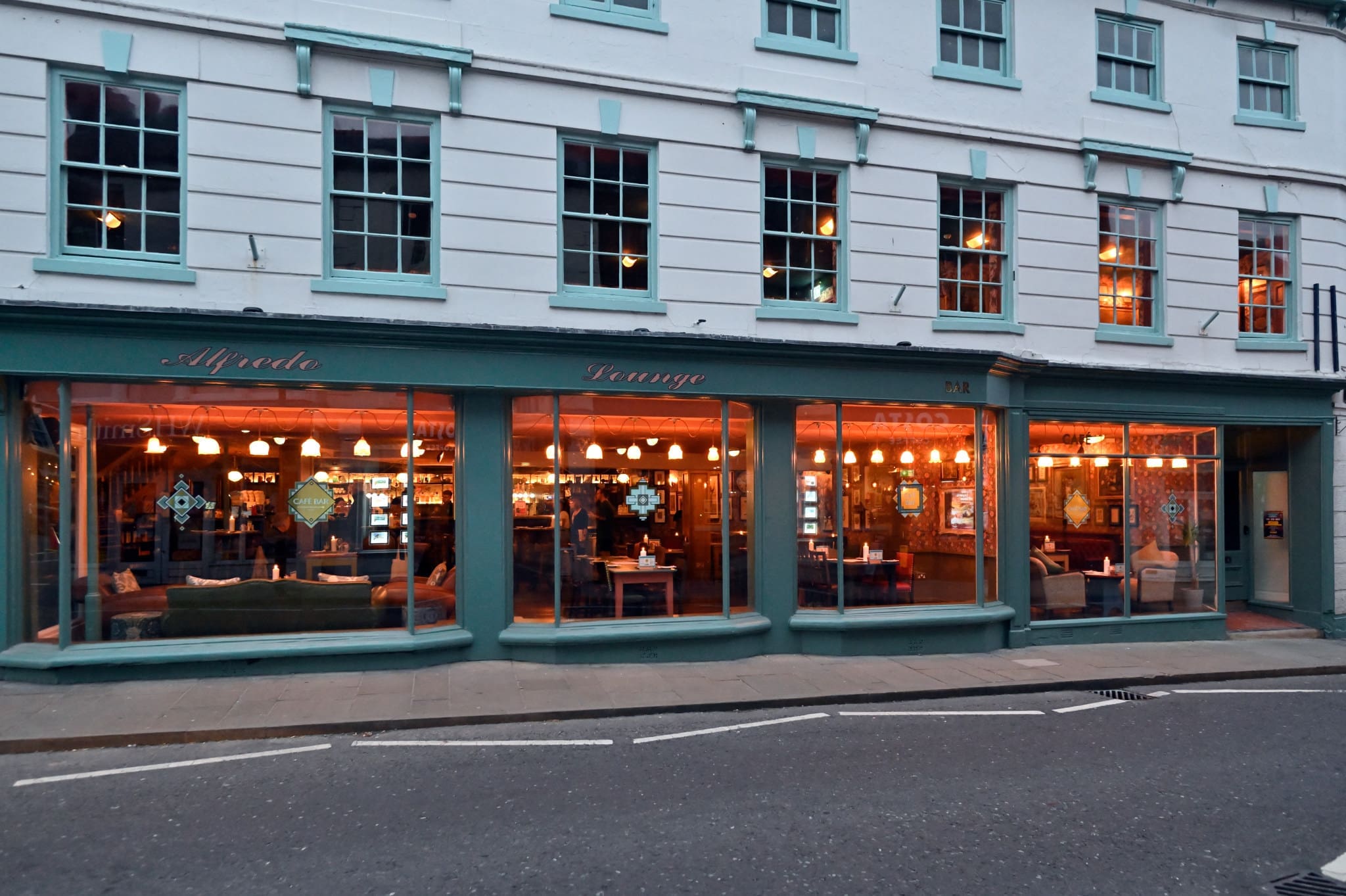 A street view of a modern lounge or cafe with large front windows, warm interior lighting, and visible seating areas. The exterior is painted teal and bordo, with signs reading "Alfredo" and "Lounge.