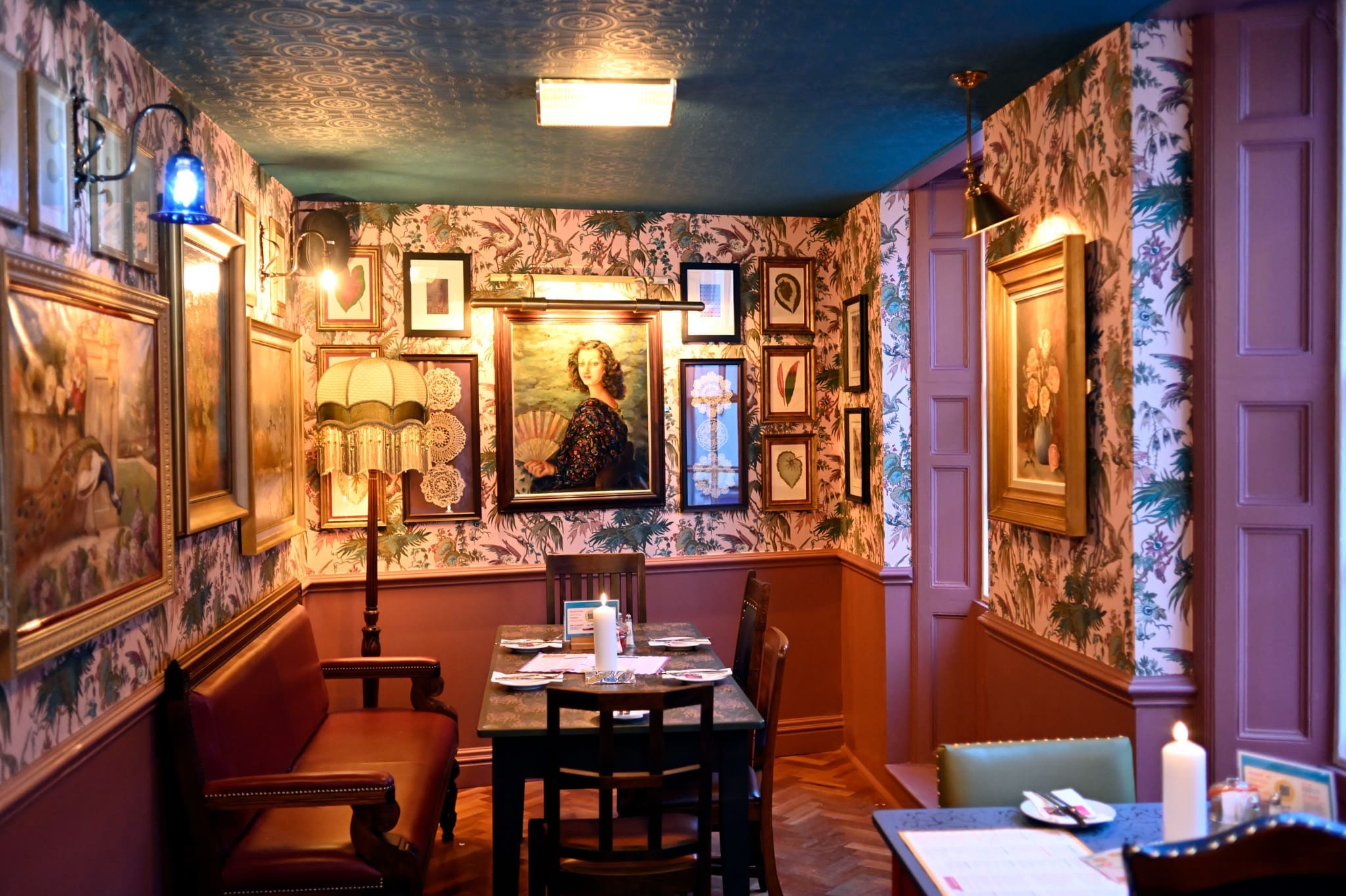 A cozy restaurant dining nook with vintage decor, floral wallpaper, framed paintings, a dark wood Bordo table set for four, cushioned chairs, soft lighting, and a patterned ceiling.