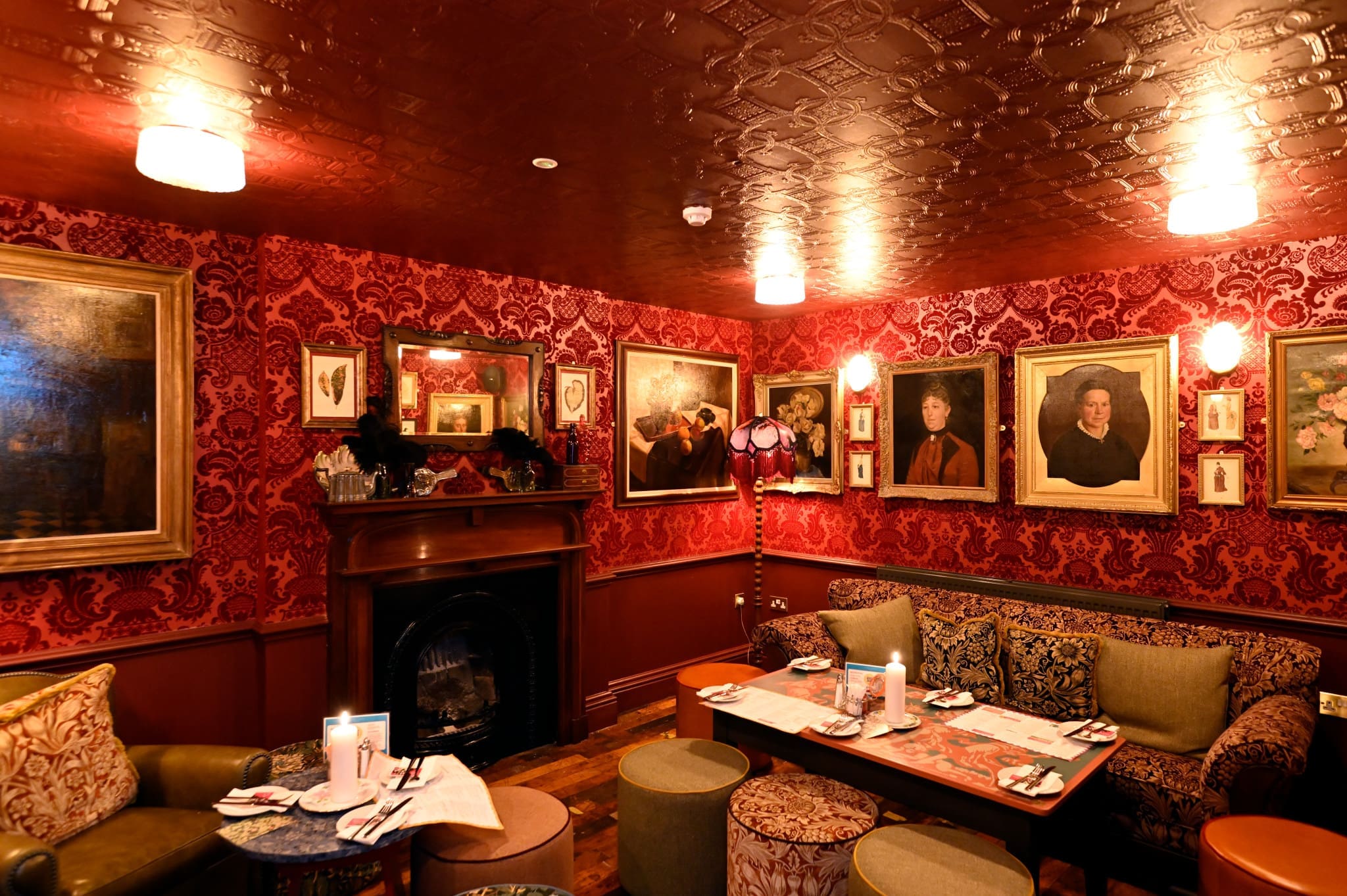 A cozy, dimly lit lounge with ornate bordo wallpaper, vintage sofas, small tables, and classic oil portraits in gilded frames. A black fireplace sits beneath a mantle, creating a warm, inviting atmosphere.