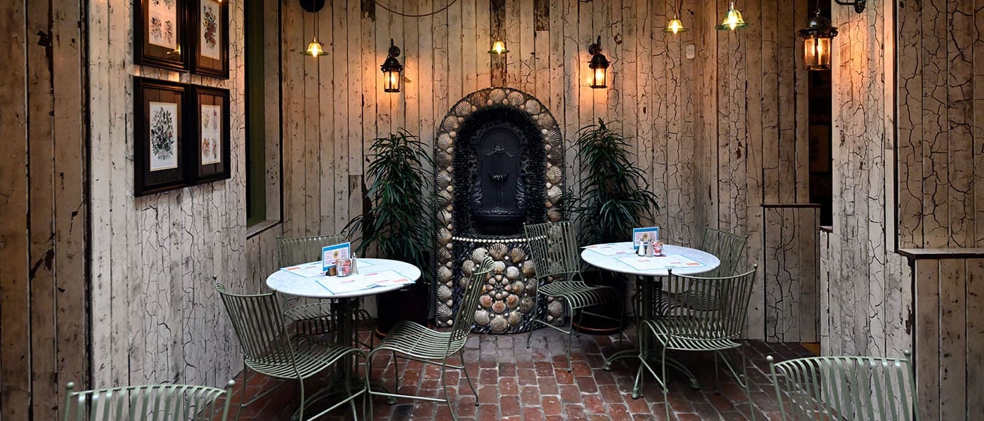 Alturo’s cozy restaurant patio features brick flooring, metal chairs, and round tables set for dining. Rustic wooden walls display framed art and a decorative stone fountain, all softly lit by elegant wall lanterns above.