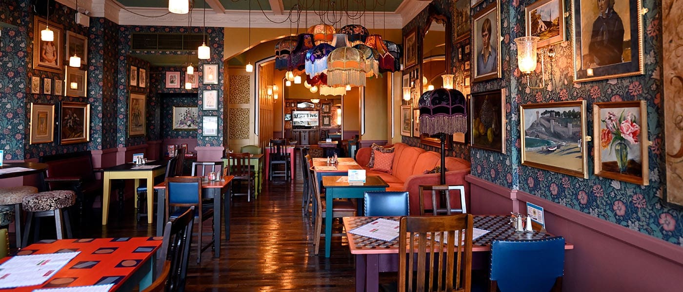 Cozy, eclectic restaurant interior at Alturo with vintage decor, floral wallpaper, mismatched chairs, framed art, colorful lampshades, and wooden floors. Tables are set for dining, creating a warm, inviting atmosphere.