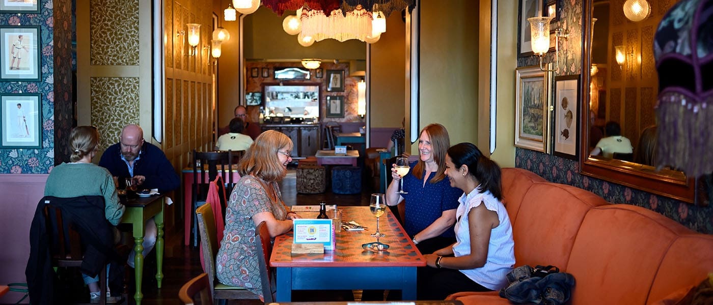 Three women sit at a colorful Alturo table in a cozy, warmly lit café, smiling and talking over drinks. Other patrons are seated at tables in the background, surrounded by decorative art and vintage wallpaper.