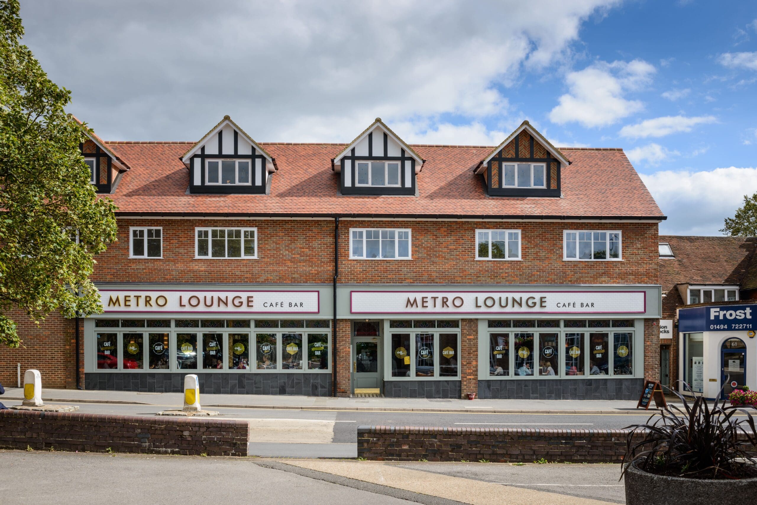 A brick building with gabled roofs houses the Metro Lounge café bar, featuring large windows with colorful Metro decorations. A tree is on the left, and a small shop named Frost is visible on the right.