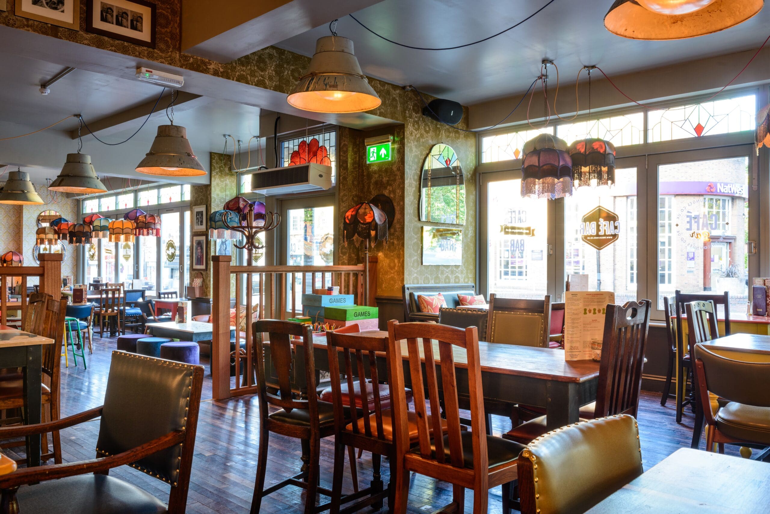 A cozy, well-lit pub interior with wooden tables and chairs, colorful lampshades hanging from the ceiling, and large windows letting in Metro views and natural light. Menus and decorations are visible throughout the space.