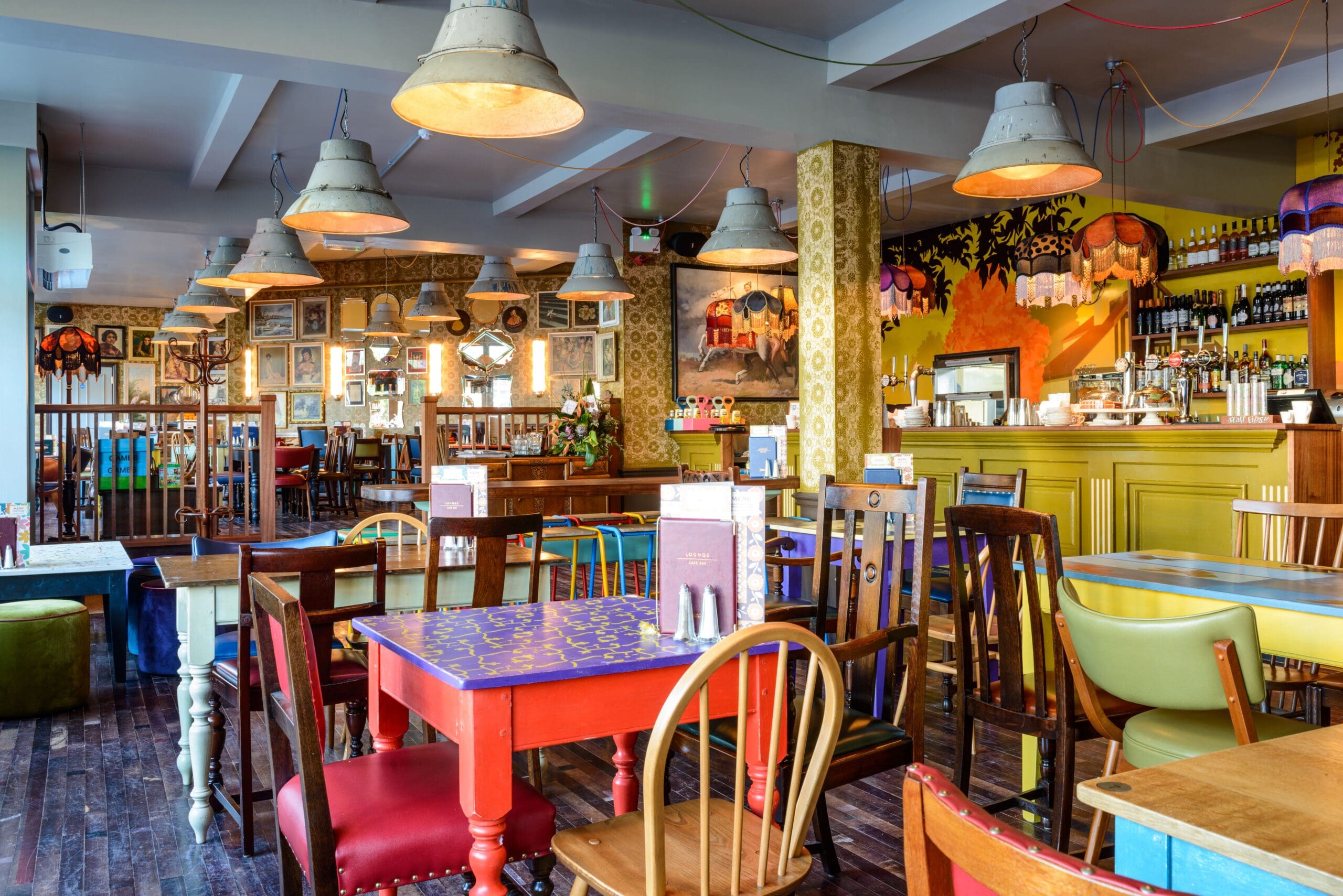Colorful, eclectic café interior with mismatched wooden chairs and tables, hanging vintage lamps, vibrant wallpaper, framed artworks on the walls, and a yellow bar area reminiscent of a lively Metro station displaying bottles and glasses.