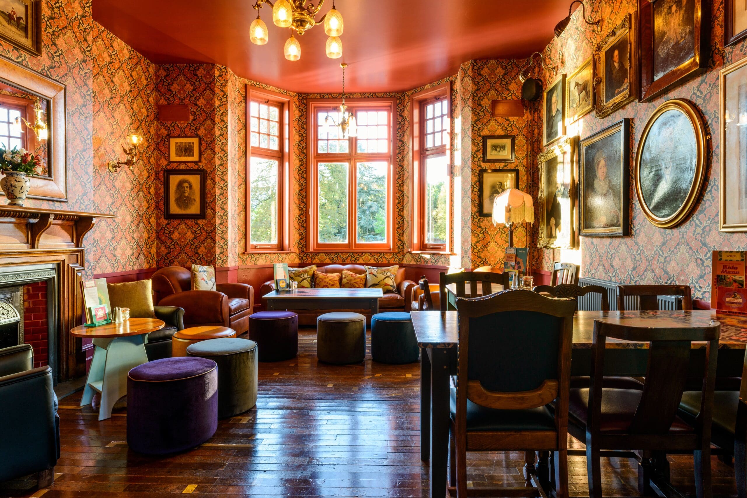 A cozy, vintage-style sitting room with colorful wallpaper, paintings on the walls, large bay windows, various chairs and sofas like the elegant Albero, small tables, and soft lighting creates a warm, inviting atmosphere.