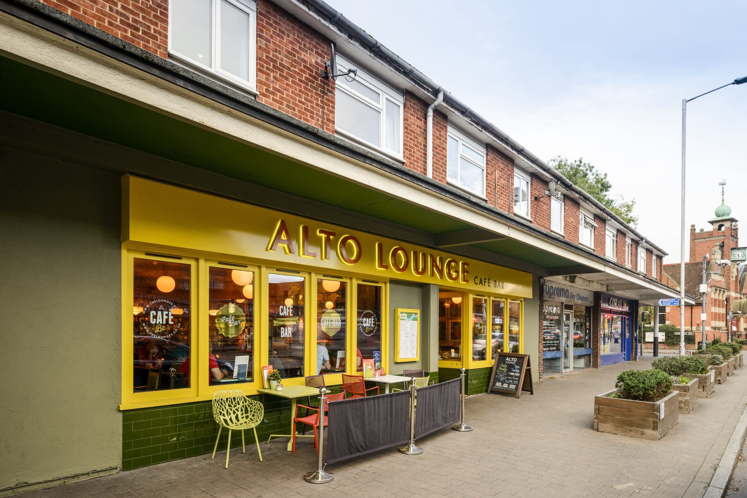 A street view of Alto Lounge, a vibrant café with a yellow exterior, large windows, and outdoor seating. The prominent Alto sign hangs above the entrance, while neighboring shops line the sidewalk under a clear sky.