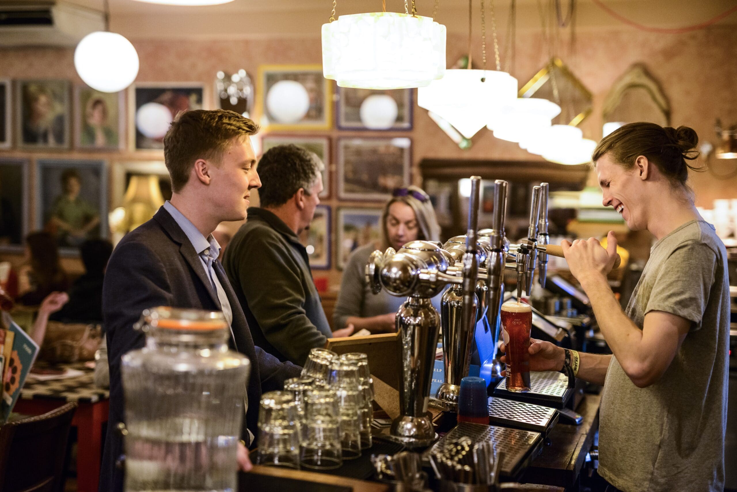 A bartender with a ponytail smiles while serving a pint of Alto beer to a customer in a suit at a cozy, warmly-lit bar. Other patrons are chatting, and framed portraits decorate the walls in the background.