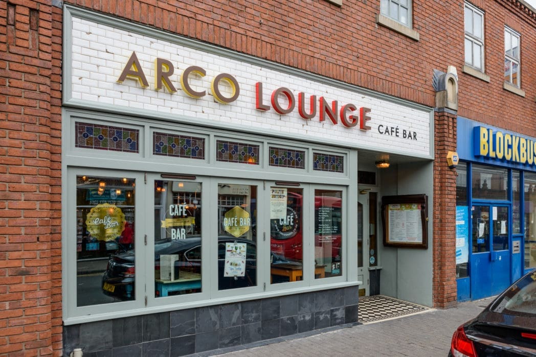 Street view of Arco Lounge Café Bar, featuring large front windows and a white Arco sign with gold and red lettering, situated next to a blue Blockbuster storefront.