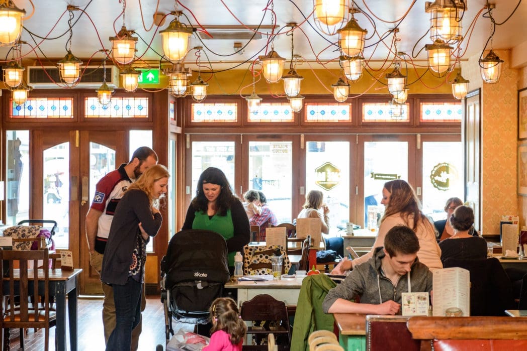 People dine and socialize in a cozy, well-lit café with Arco hanging lights and large windows. Some guests are seated, reading menus, while others stand near a stroller and talk. The atmosphere is lively and inviting.