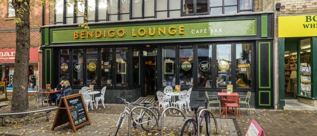 Street view of Bendigo Lounge café bar with outdoor seating, bicycles parked in front, and colorful Bendigo storefronts on either side; autumn leaves are scattered on the ground.