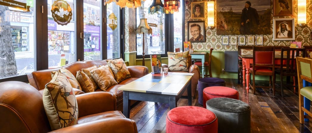 A cozy, colorful lounge in Bendigo with leather sofas, patterned cushions, round velvet stools, eclectic hanging lamps, and vintage decor. Large windows overlook a lively street with shops. Framed photos and portraits cover the walls.