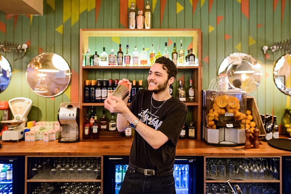 A smiling bartender with a beard and curly hair shakes a cocktail shaker behind a colorful, well-stocked Bendigo bar, with bottles, glasses, and jars of fruit on display.