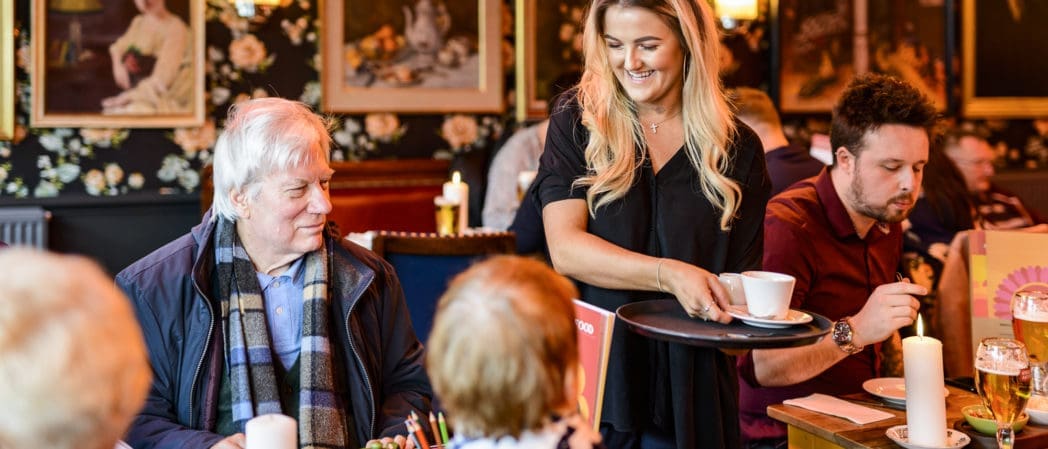A smiling waitress serves coffee to an older man, Benito, sitting with others at a warmly lit restaurant, while another man nearby eats. The background features floral wallpaper and framed portraits.