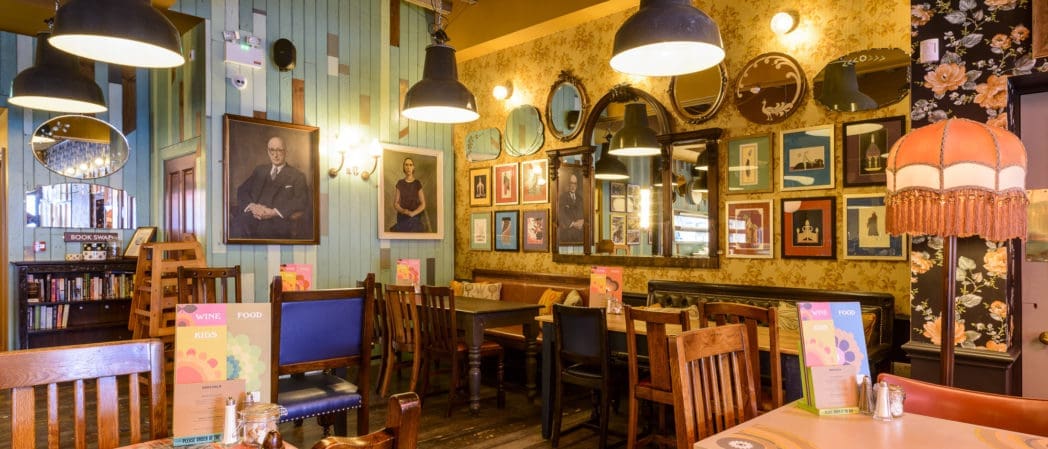 Cozy restaurant interior with wooden tables and chairs, decorative mirrors and framed portraits of Benito on colorful walls, hanging pendant lights, bookshelves, and menus on each table. Warm, inviting atmosphere with vintage decor.