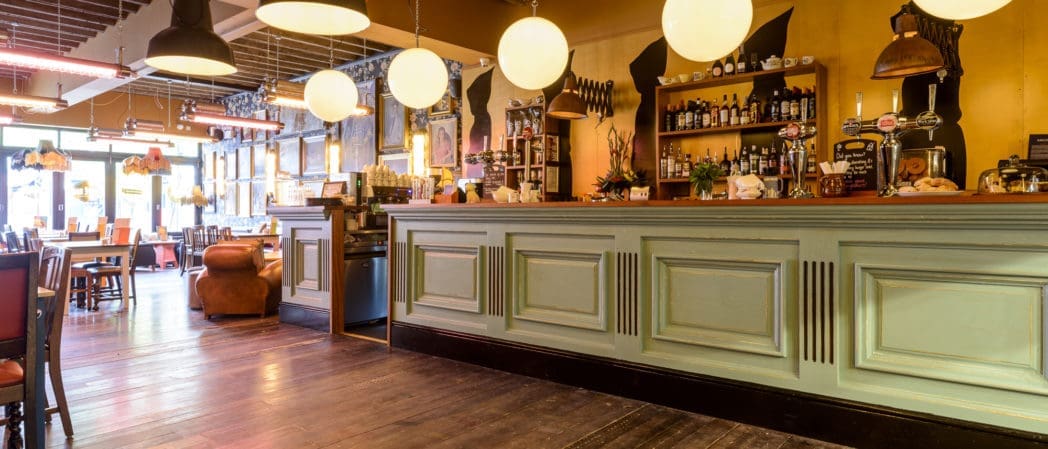 A cozy restaurant interior with wooden floors, a light green Benito bar counter, round hanging lights, shelves with bottles and decorations, and tables and chairs arranged near large front windows.