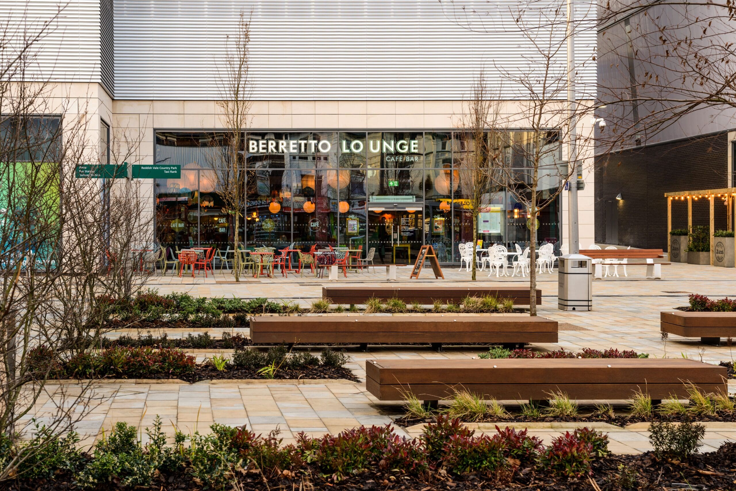 Outdoor view of Berretto Lounge, a modern café with a glass front, colorful chairs and tables outside, and landscaped planters with small shrubs in the foreground.